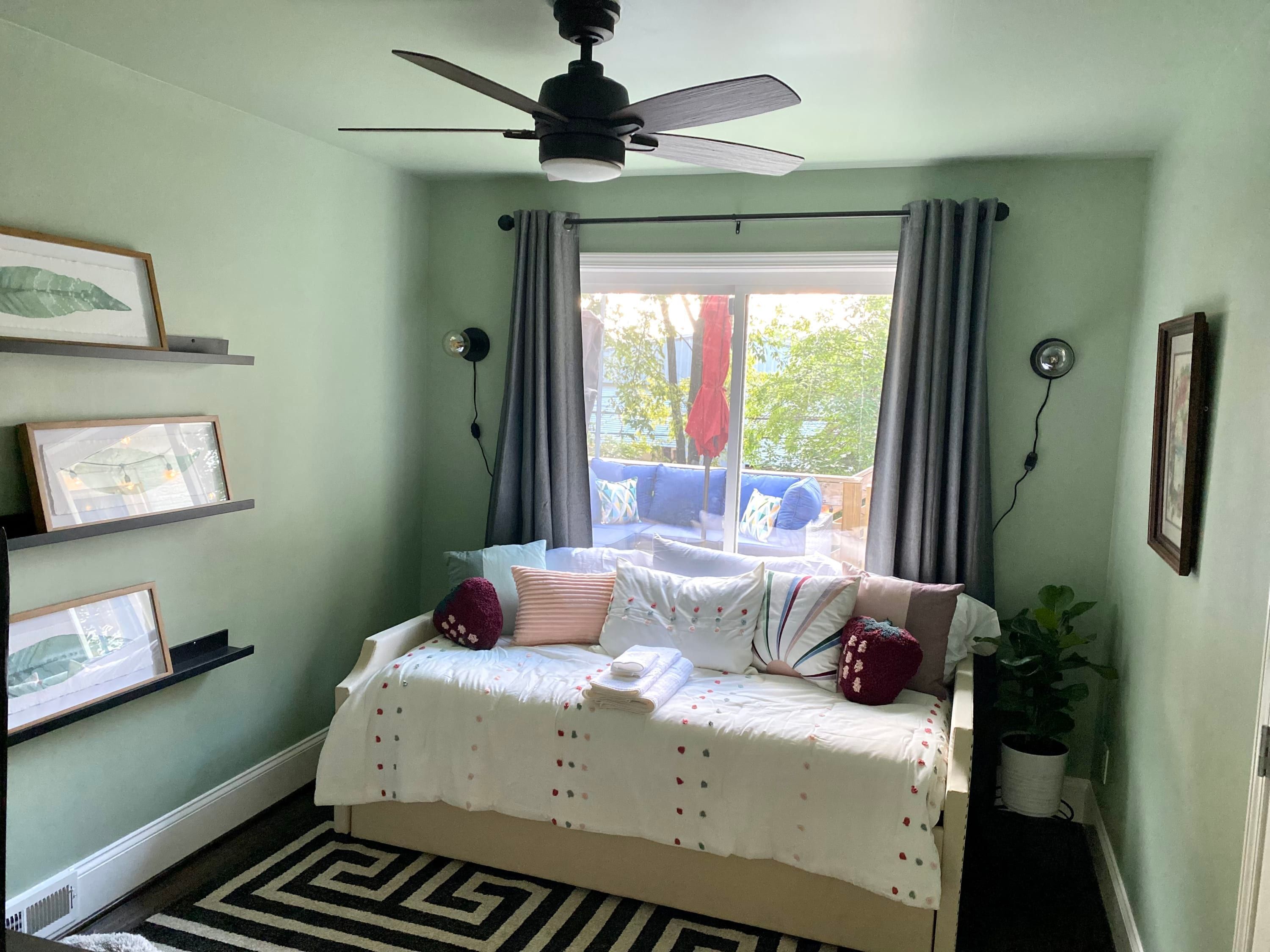 Ein Schlafzimmer mit einem Bett, einem Deckenventilator, einem Fenster mit Vorhängen und gerahmten Bildern an der Wand.