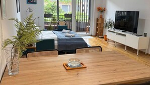 Een moderne woonkamer met een houten eettafel, twee zwarte stoelen, een bank, een televisie en een groot raam met uitzicht op de omgeving.