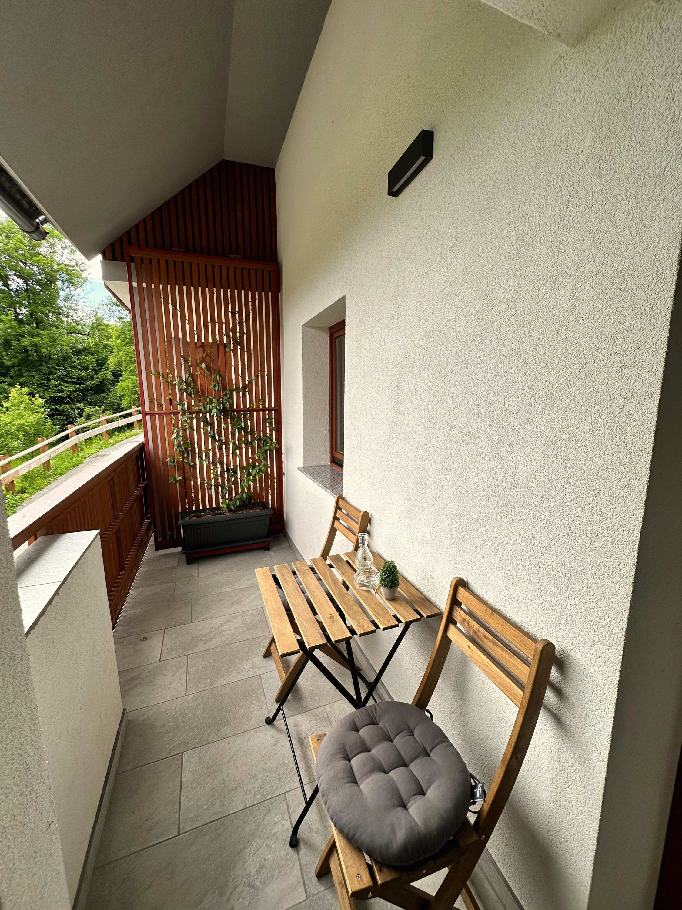 A small balcony with a wooden table and chairs, a potted plant, and a view of greenery.