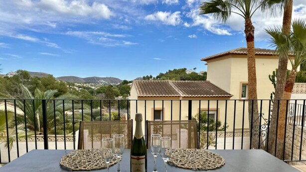 A table set with a bottle of wine and glasses, overlooking a scenic landscape with palm trees and a building in the background. - Three Bedroom Villa With Private Pool in Moraira (Teulada)