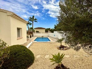 Una piscina rodeada de grava, un pequeño jardín, un edificio blanco y una palmera.