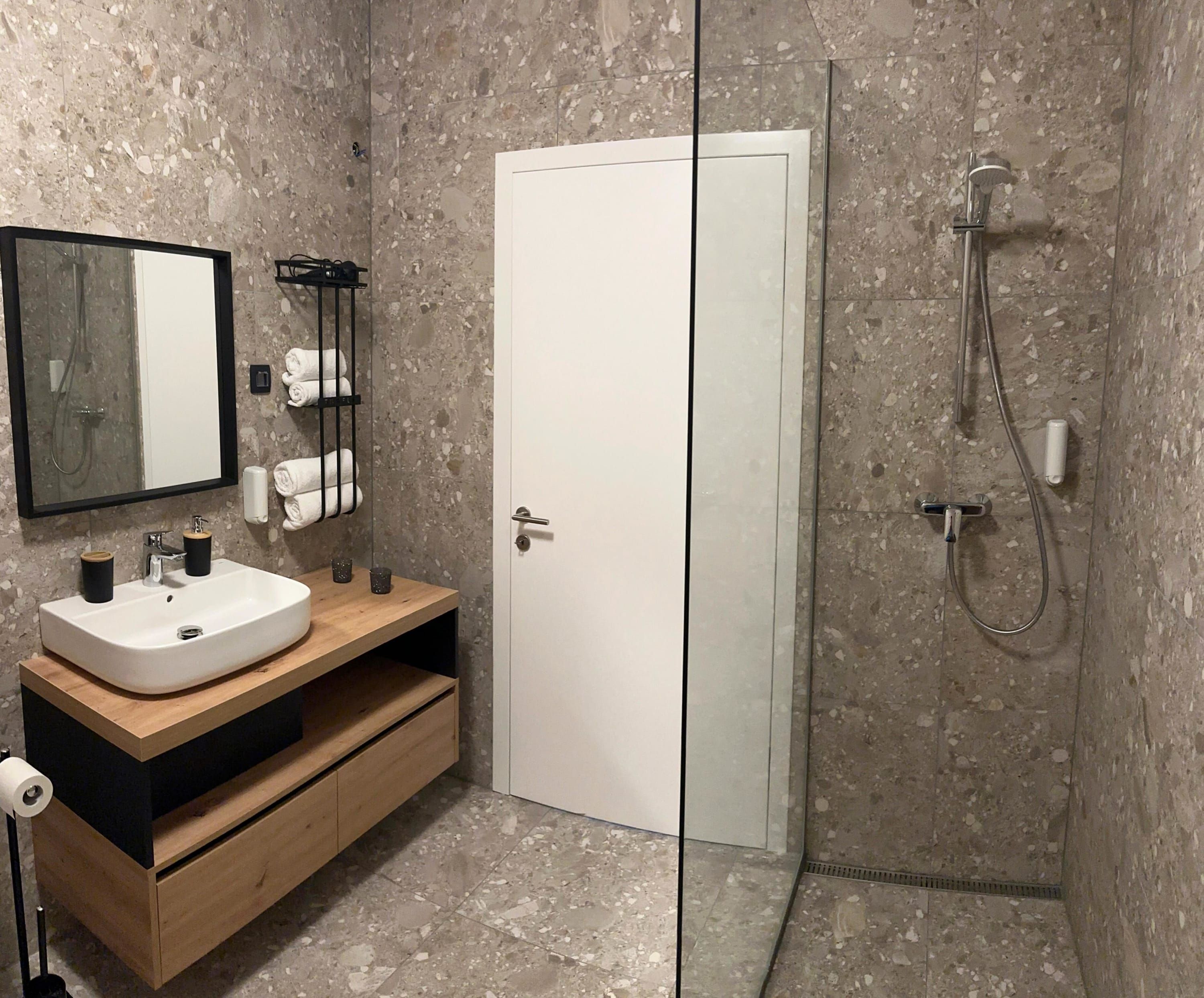 Un baño moderno con un lavamanos blanco, tocador de madera, cabina de ducha de vidrio y suelo de baldosas de mármol.
