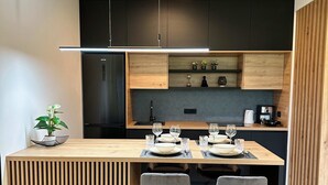 A modern kitchen with a central island, dark cabinetry, and a dining area with a grey sofa and bar stools. - Soca Valley Suites - Premium Apartment A15 (Bovec)