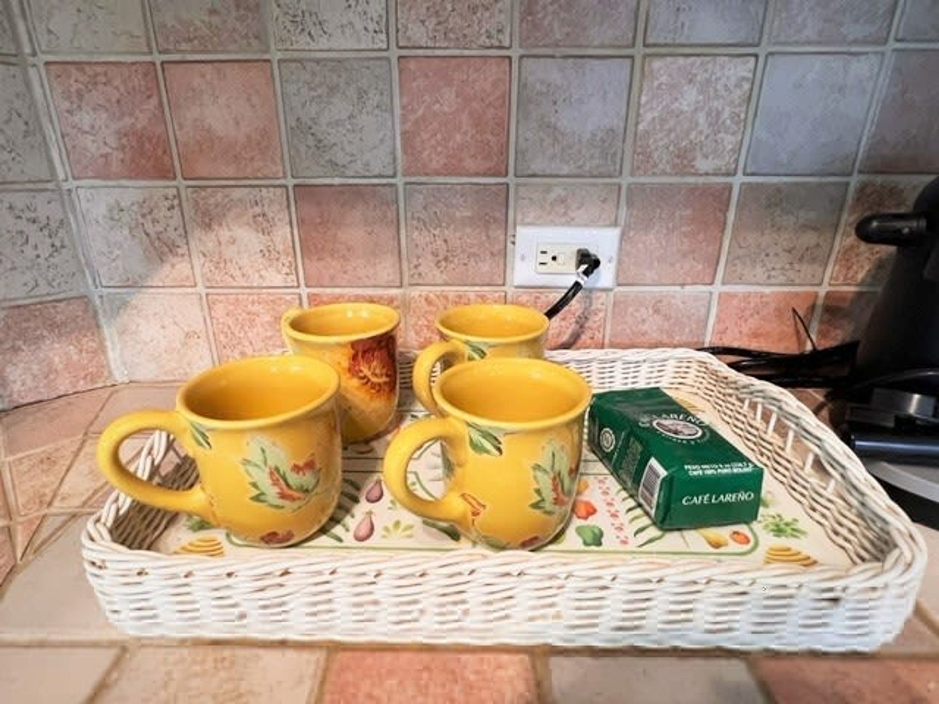 A kitchen counter with a wicker tray holding four yellow mugs and a box of Café Laredo coffee.
