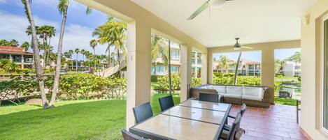 A covered patio with a table and chairs, a ceiling fan, and a view of a landscaped area with palm trees and buildings in the background.