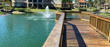 A wooden boardwalk leading to a body of water with a fountain, surrounded by palm trees and buildings with red roofs.