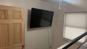 A room with a wooden door, a mounted flat-screen TV, a ceiling fan, and a window with blinds.