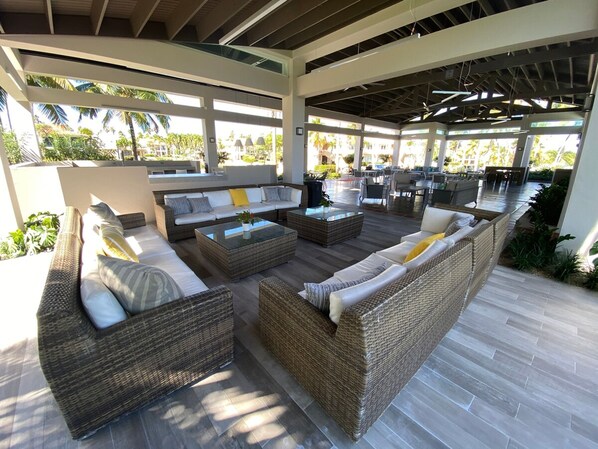 A spacious outdoor lounge area with wicker furniture and a glass coffee table, featuring a view of palm trees and a pool. - Villa Quenepa at Lakeside Villas (Vega Alta)
