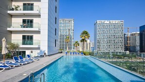 A swimming pool with lounge chairs, a modern building with balconies, and palm trees in the background. - Luxurious Studio With Bay View in Sol Bay (Dubai)