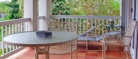 A covered outdoor seating area with a round table and chairs, surrounded by a white railing, overlooking a garden.