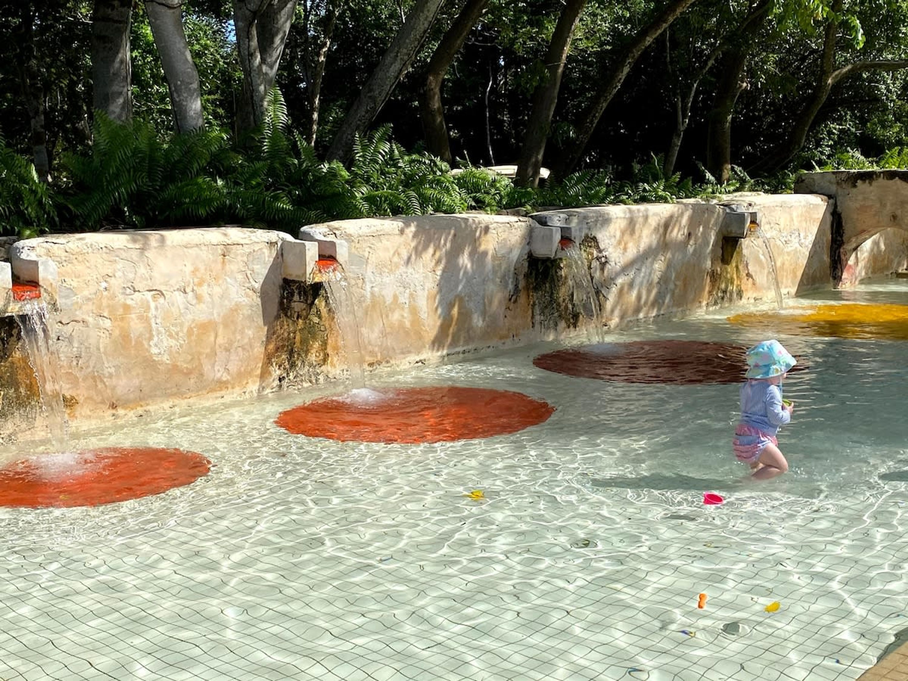 Niño jugando en una zona de juegos con agua poco profunda, suelo de rejilla y elementos acuáticos circulares.