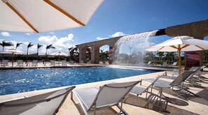 Zona de piscina con sillones de descanso y sombrillas, un elemento acuático y una estructura de piedra al fondo.