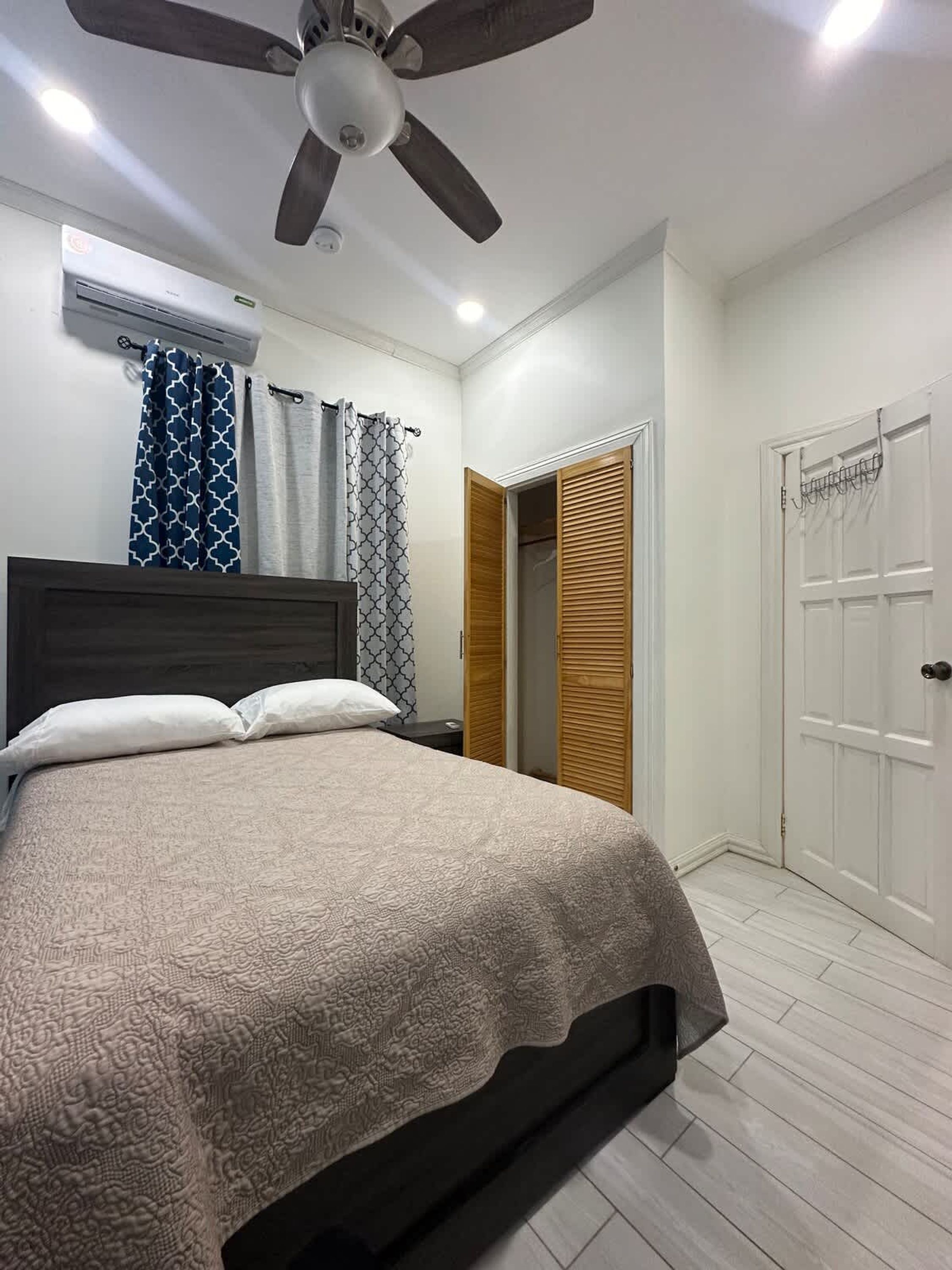 Un dormitorio con cama, ventilador de techo, aire acondicionado, cortinas estampadas y una puerta de madera.
