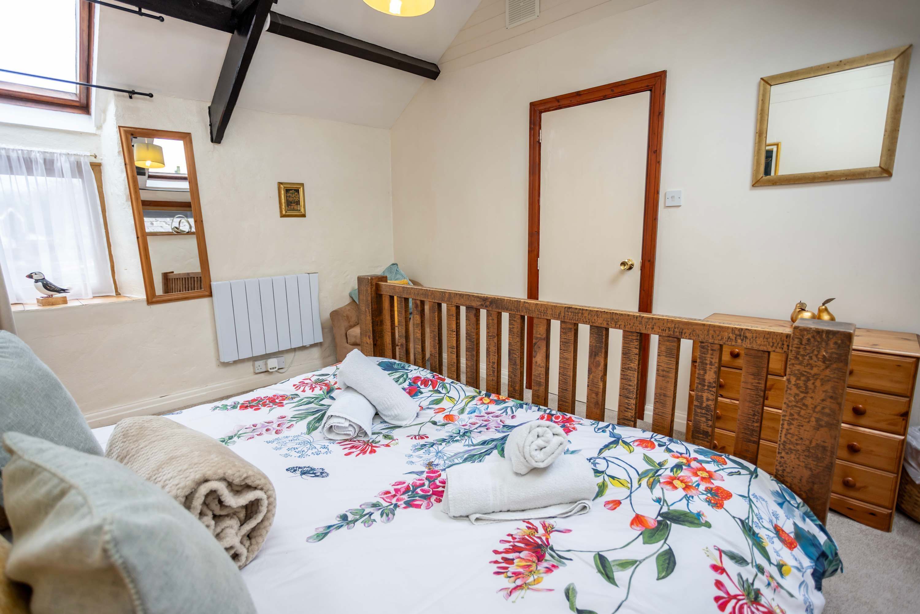 Un dormitorio con cabecera de madera, cama con ropa de cama floral, mesita de noche, cómoda, espejo y ventana con cortinas.