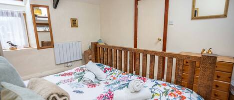 Un dormitorio con cabecera de madera, cama con ropa de cama floral, mesita de noche, cómoda, espejo y ventana con cortinas.
