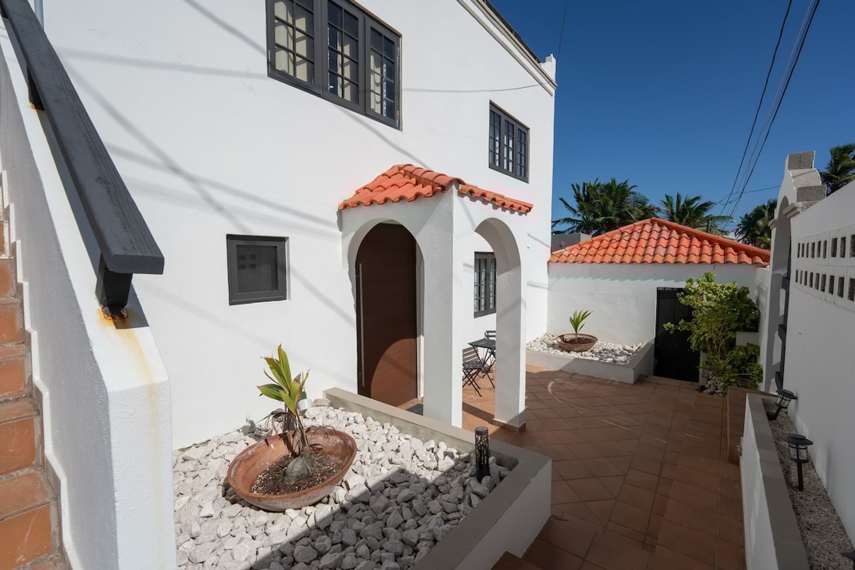 A white building with a red-tiled roof, a small patio with a potted plant, and a clear blue sky.