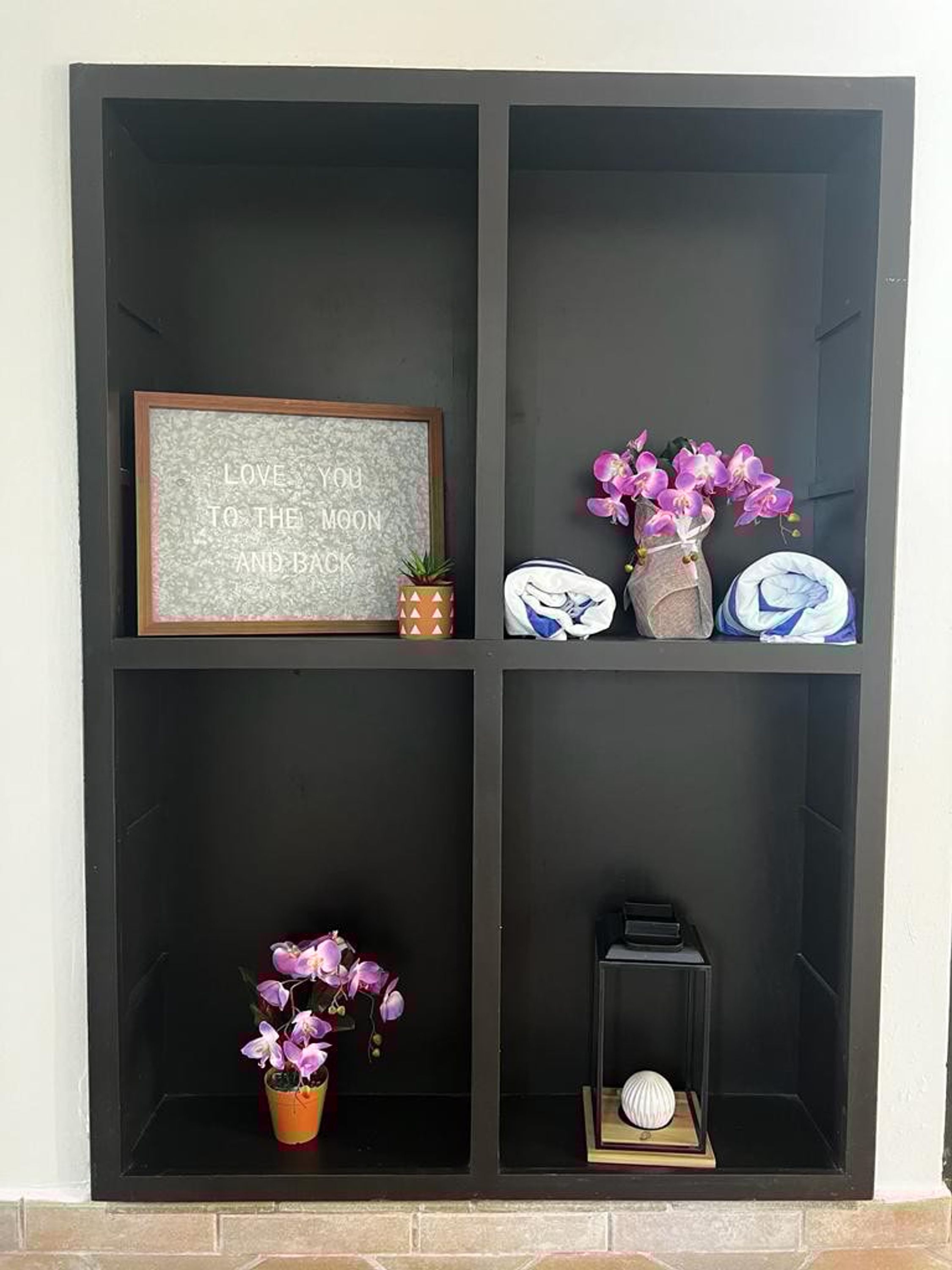 A black shelving unit with four compartments. The top left shelf holds a framed sign with text, the top right shelf has a vase with purple flowers and a pair of shoes, the bottom left shelf contains a small potted plant, and the bottom right shelf has a small decorative item.