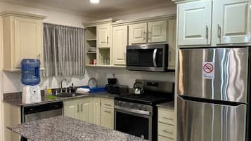 Una cocina con armarios blancos, encimera de granito, refrigerador de acero inoxidable, microondas, estufa y dispensador de agua.