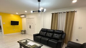 A living room with a black leather sofa, a glass coffee table, and a TV mounted on a yellow wall. - Gentlemen s Suite 3 Bedroom (Georgetown)