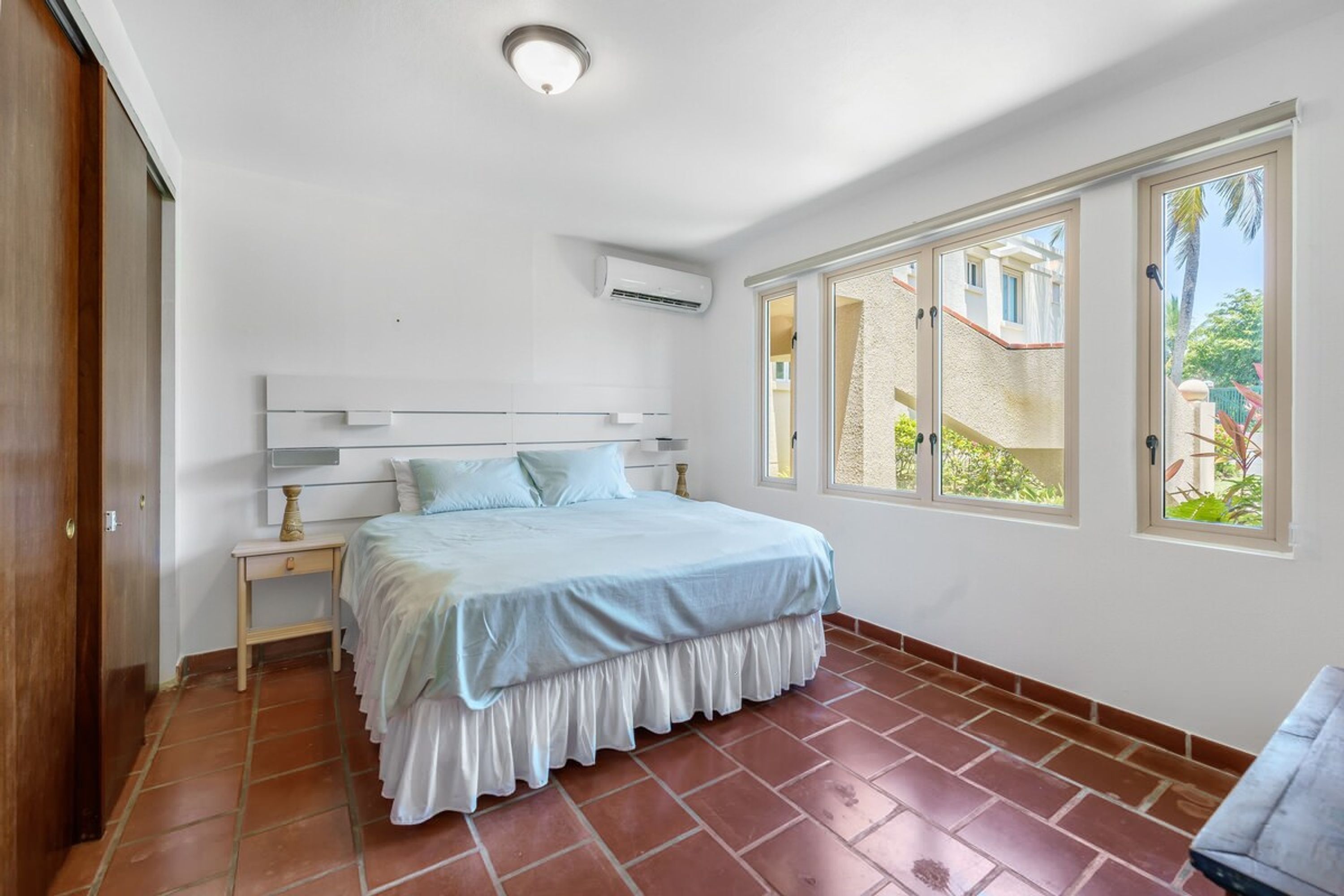Un dormitorio con cama, mesita de noche, aire acondicionado y ventana con vistas a un edificio y vegetación.