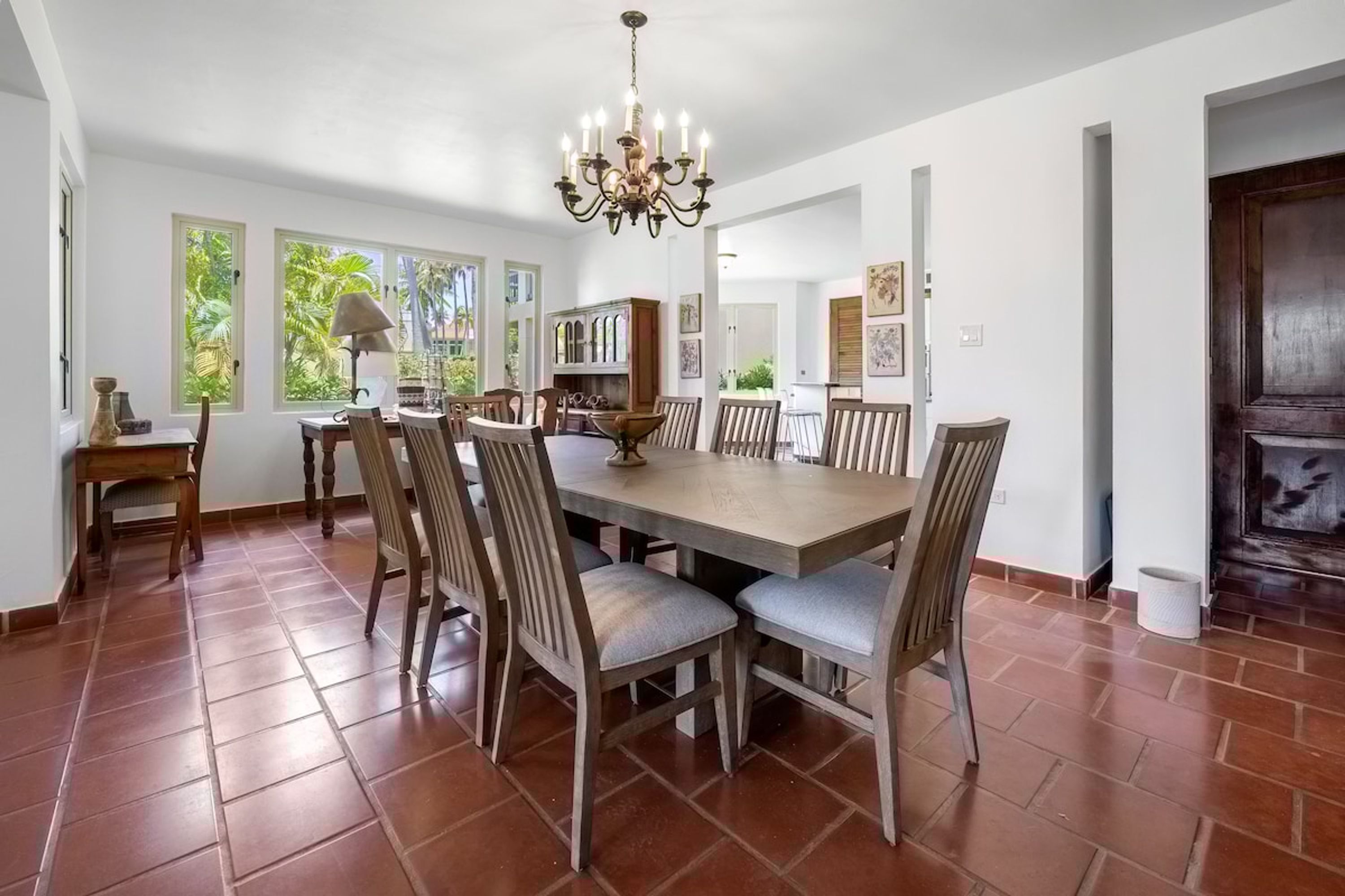 A dining area with a large wooden table, multiple chairs, a chandelier, and a view of the outdoors through large windows.