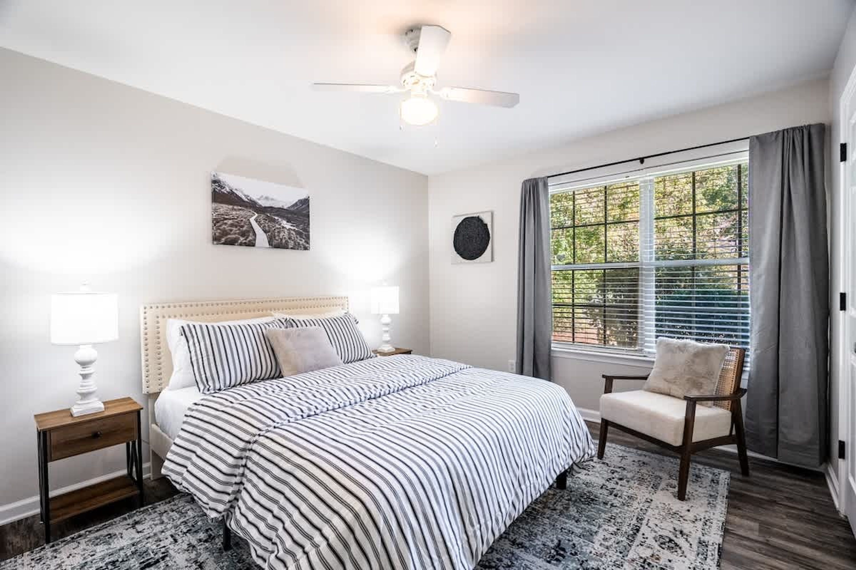 A bedroom with a bed, a chair, a nightstand, a ceiling fan, a window with curtains, and a wall-mounted picture.