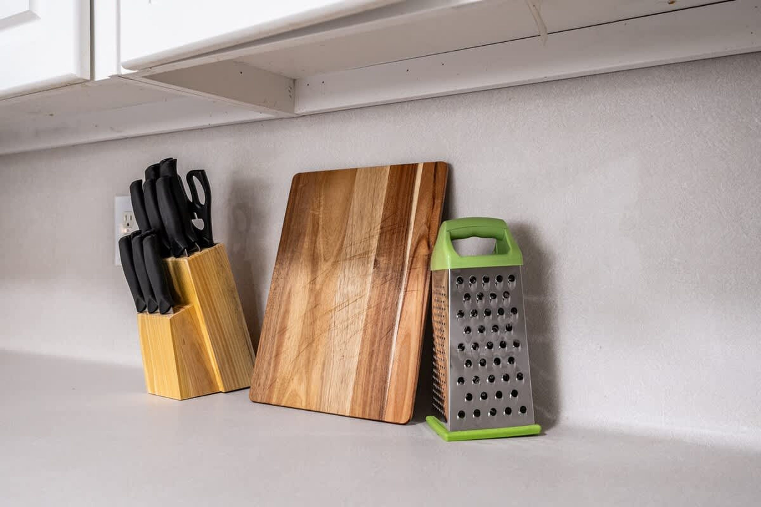 Un piano di lavoro in cucina con un tagliere di legno, un portacoltelli e una grattugia verde.