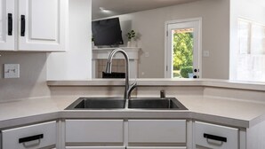 A modern kitchen with a stainless steel sink, white cabinetry, and a window with a view of greenery. - Pool Oasis w King Bed Firepit (Cabot)