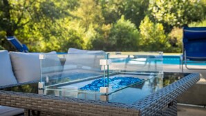 A glass-top table with a metal frame, surrounded by wicker furniture, set outdoors with a pool and trees in the background. - Pool Oasis w King Bed Firepit (Cabot)