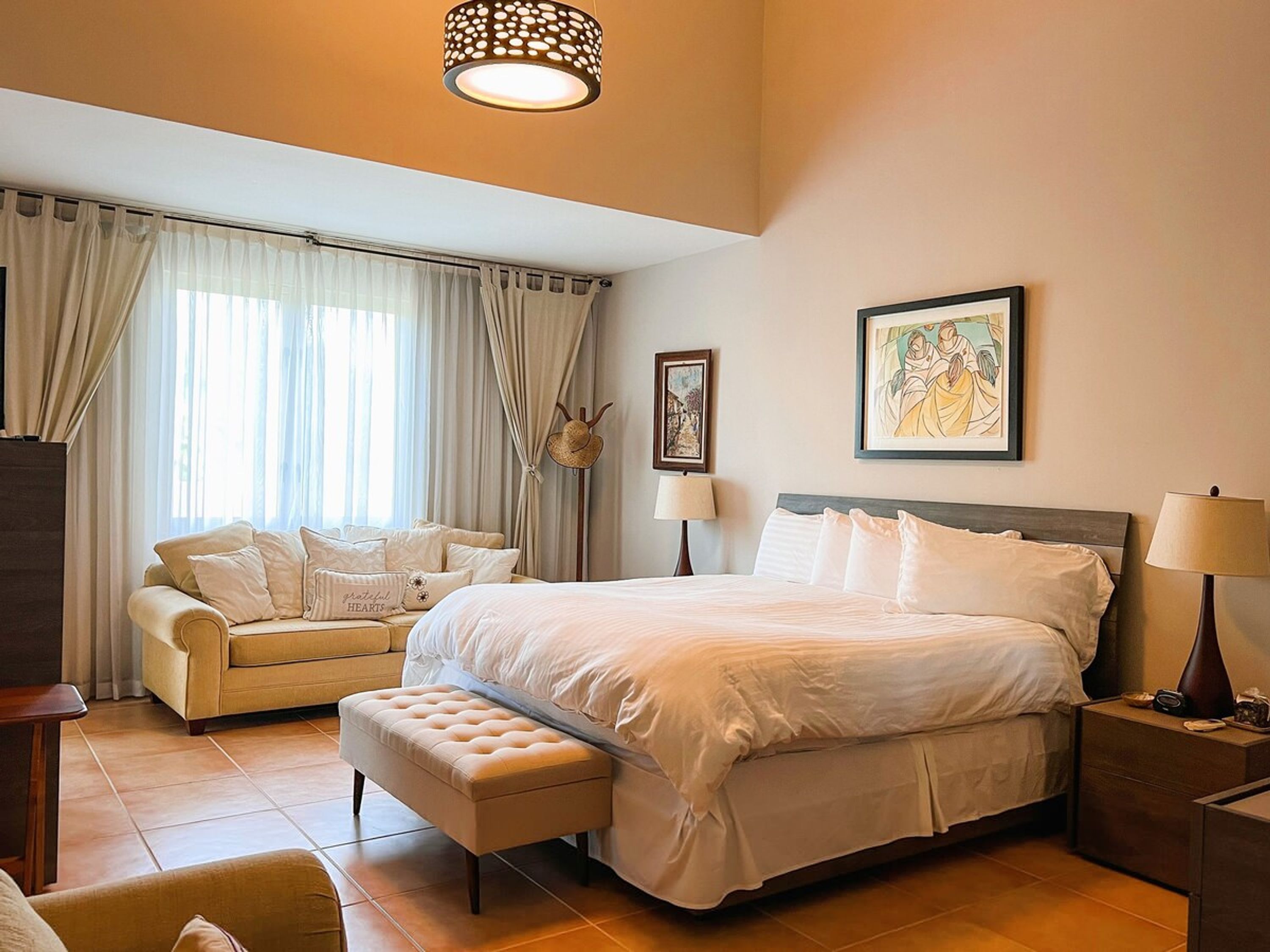A hotel room with a bed, a sofa, a bench, a lamp, a picture on the wall, and a window with curtains.