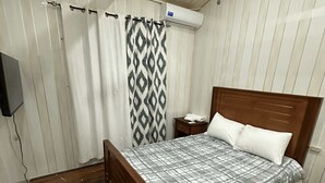 Ένα υπνοδωμάτιο με ένα ξύλινο κρεβάτι, ένα γκρι και λευκό καρό σεντόνι, μια διακοσμητική κουρτίνα για το ντους και έναν ανεμιστήρα οροφής.