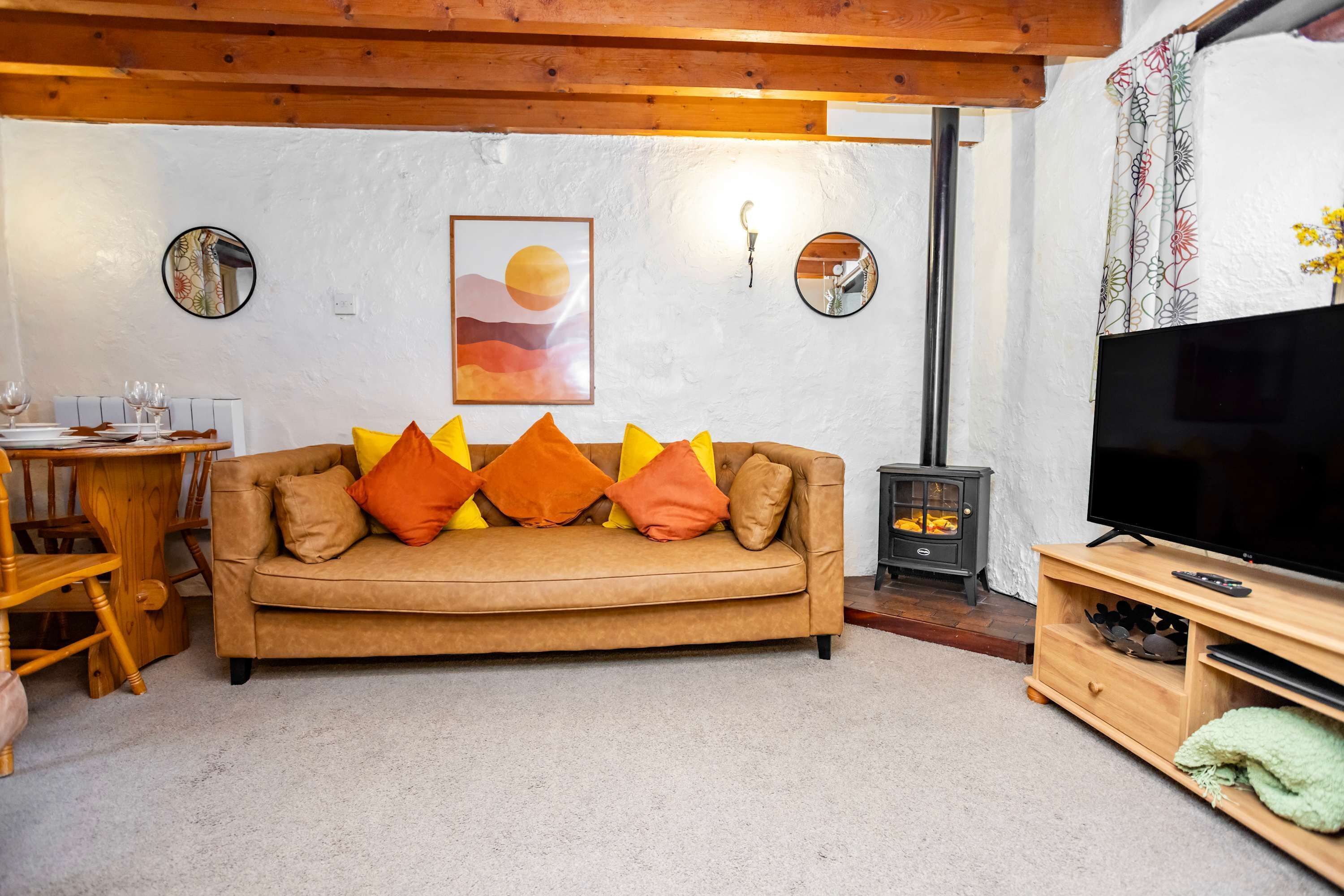Una sala de estar acogedora con un sofá marrón, cojines de colores, un mueble de TV de madera, una chimenea y un comedor con mesa y sillas de madera.