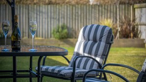 A patio area with a metal table, two wine glasses, and a bottle of wine. There is a striped outdoor chair and a cushioned bench. - Johnnys Cove - 2 Bedroom House - New Hedges (Saundersfoot)