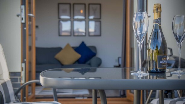 A glass table with a bottle of Nozema Champagne and two glasses, a striped chair, and a sofa with blue and yellow cushions in the background. - Johnnys Cove - 2 Bedroom House - New Hedges (Saundersfoot)