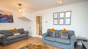 A living room with two grey sofas, wooden coffee tables, and a cityscape painting. - Johnnys Cove - 2 Bedroom House - New Hedges (Saundersfoot)