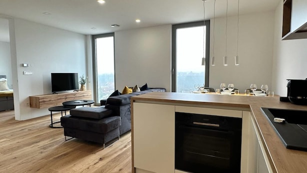 A modern kitchen with a built-in oven, a dining area with a wooden table and chairs, a dark-colored sofa, and a large window providing a view of the city.
