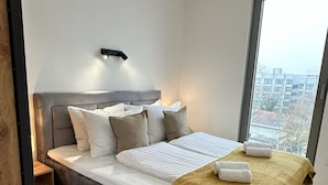 A modern bedroom with a large bed, a nightstand, a wall-mounted light, and a large window offering a city view.