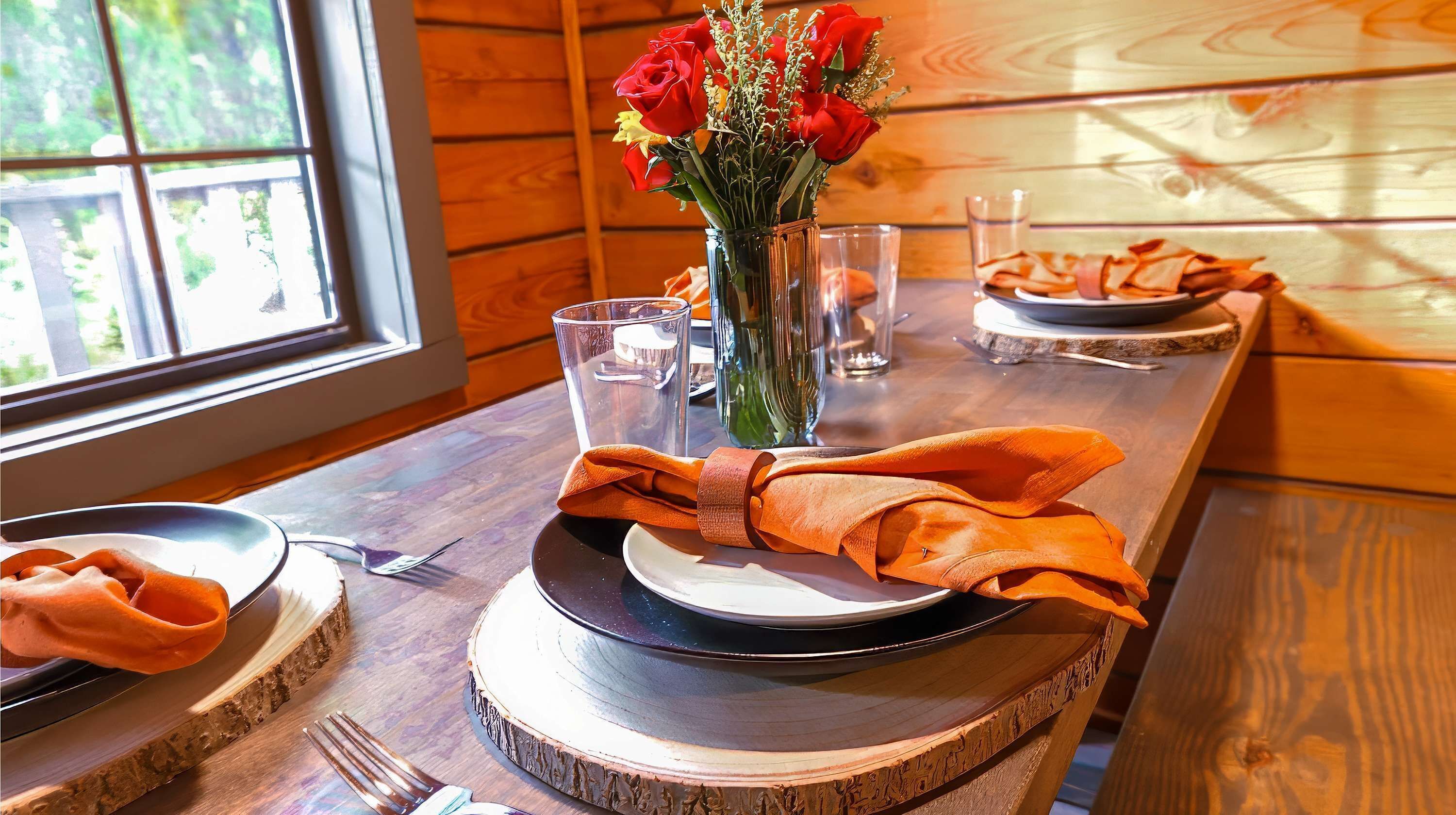 A wooden dining table with a vase of red flowers, clear glasses, and neatly folded orange napkins.