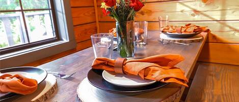 A wooden dining table with a vase of red flowers, clear glasses, and neatly folded orange napkins.