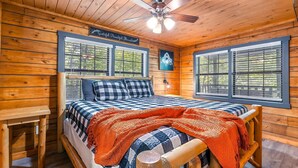 A wooden cabin bedroom with a bed, two windows, a ceiling fan, and a sign above the bed.