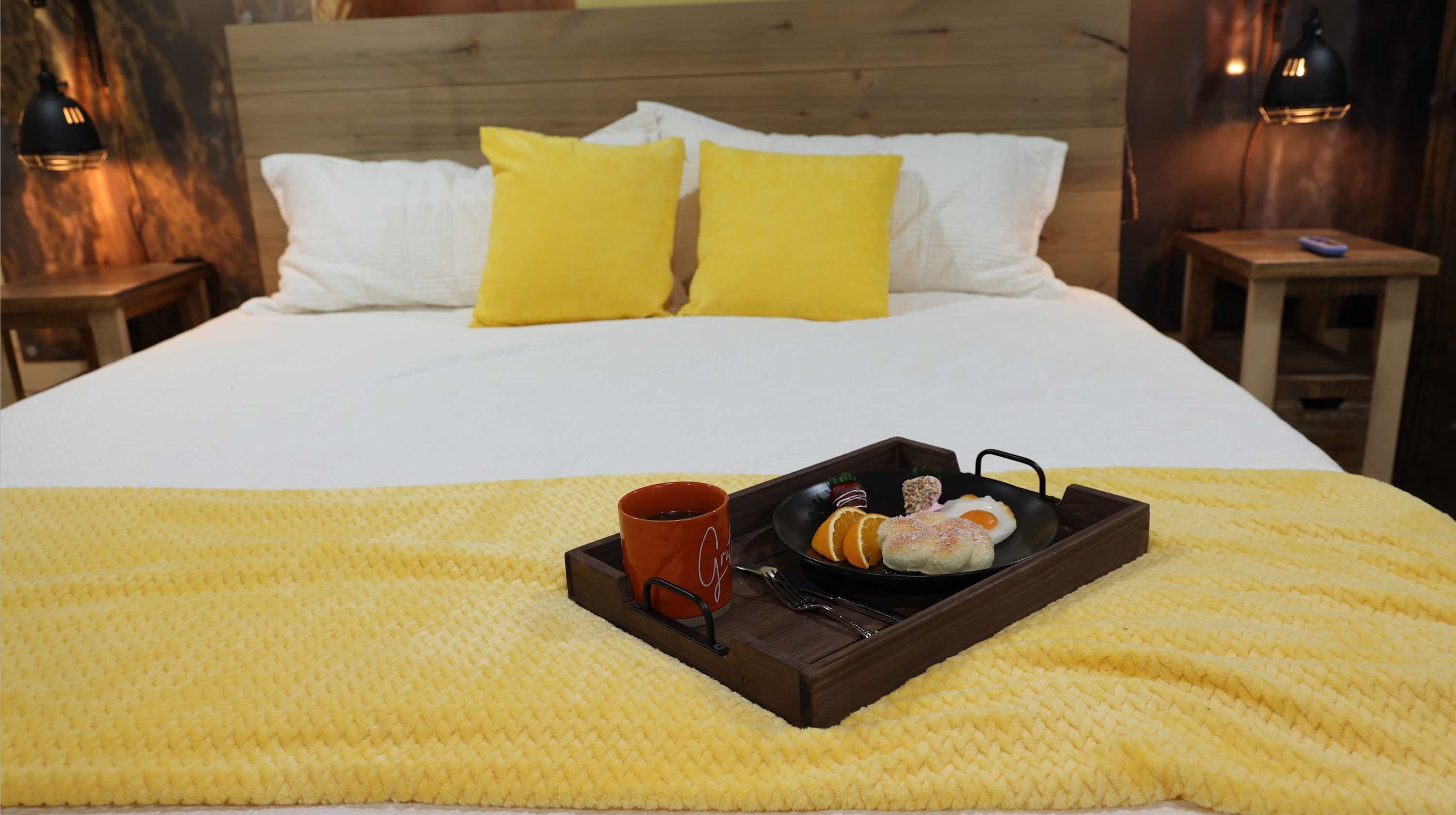 Una cama con ropa de cama blanca, almohadas amarillas, una bandeja con comida y una taza, y dos lámparas de noche.
