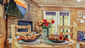 L’intérieur d’un chalet en bois, avec une table à manger dressée pour deux personnes, une cheminée et un tableau représentant un coucher de soleil.