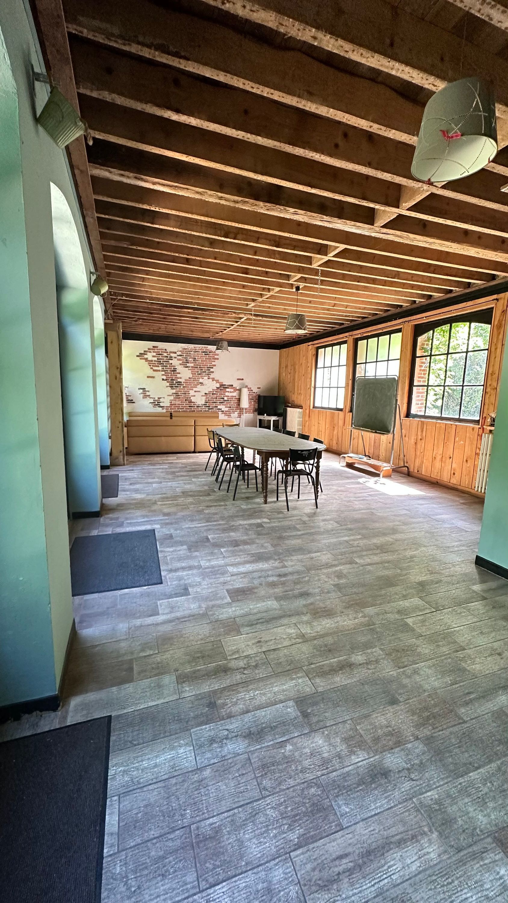 A spacious room with a large wooden table and chairs, a brick wall mural, and large windows.