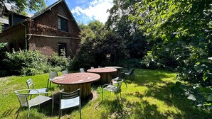 A rustic outdoor seating area with round wooden tables and metal chairs, surrounded by lush greenery and a brick building. - Ancienne Orangerie Du Chateau (Les Hauts-de-Caux)