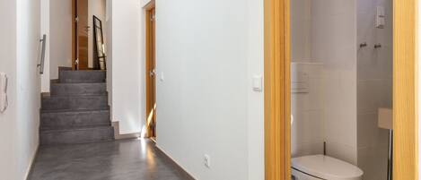 Un interior moderno con piso de concreto gris, una puerta de madera que da al baño y una escalera al fondo.