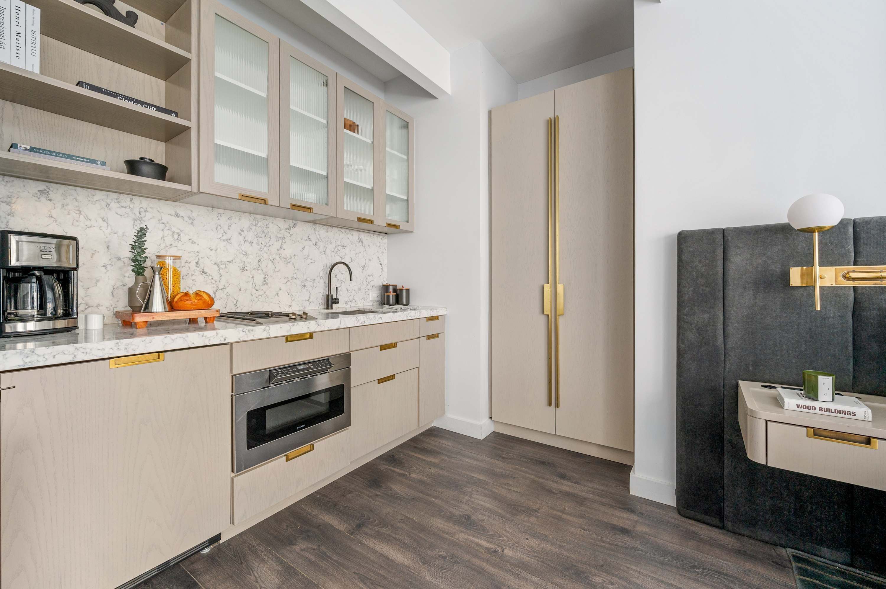 A modern kitchen with a marble countertop, stainless steel appliances, and a built-in oven. There are wooden cabinets with gold handles and a large cabinet with glass doors.