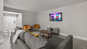 Un salon moderne avec un canapé gris, deux fauteuils orange, une table basse en bois et un tél éviseur fixé au mur qui affiche des services de diffusion en continu.