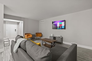 A modern living room with a grey sofa, two orange chairs, a wooden coffee table, and a mounted TV displaying streaming services. - Welcoming 2BR 2BA Retreat With Modern Comforts (Rochester)