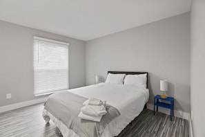 A neatly made bed with a white comforter and a gray throw blanket, a blue nightstand with a white lamp, and a window with closed blinds. - Welcoming 2BR 2BA Retreat With Modern Comforts (Rochester)