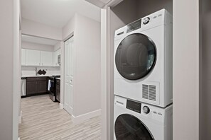 A laundry room with stacked washers and dryers, a kitchen area with cabinets and appliances, and a hallway leading to a door.